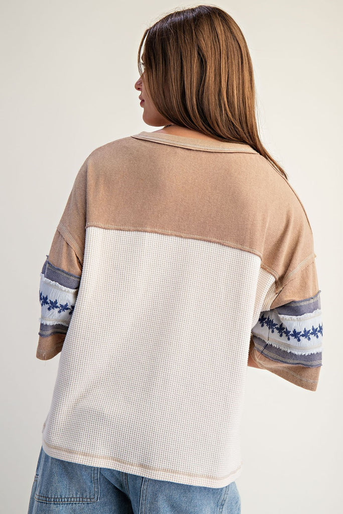 Model wearing a snow-washed waffle knit varsity t-shirt with beige jersey sleeves, large number 12 graphic in blue at front, and embroidered floral detailing on the wide short sleeves. Styled with relaxed denim bottoms.