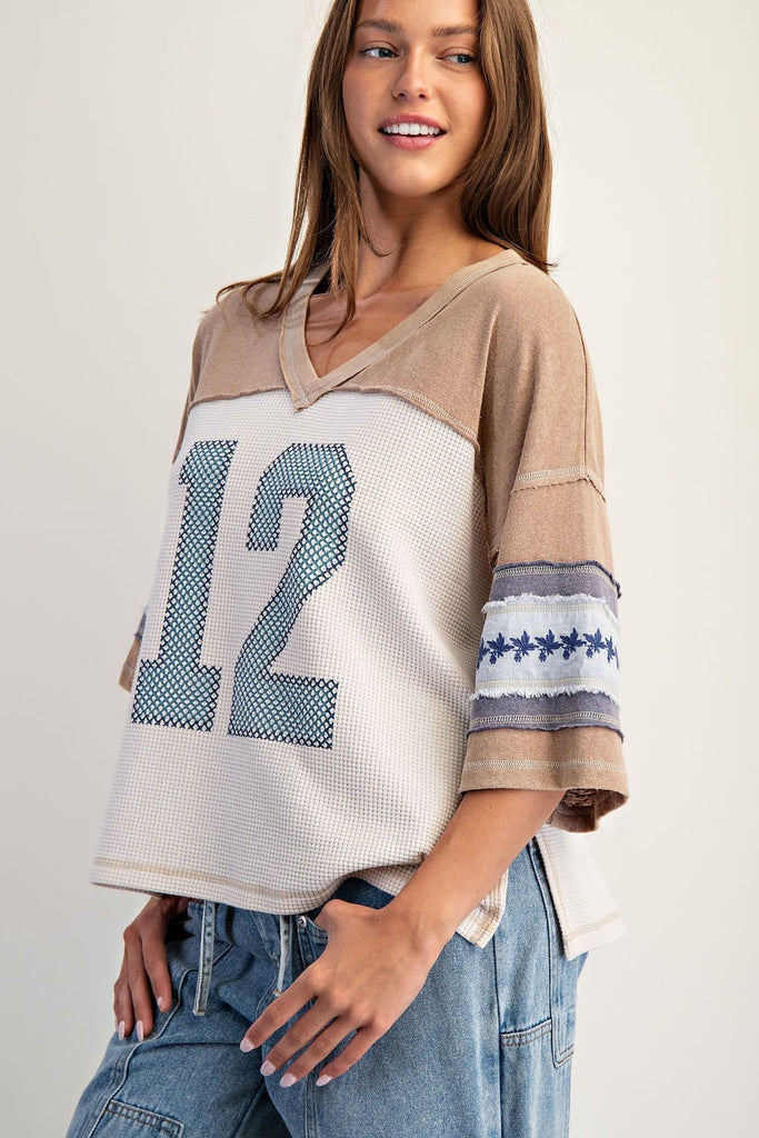 Model wearing a snow-washed waffle knit varsity t-shirt with beige jersey sleeves, large number 12 graphic in blue at front, and embroidered floral detailing on the wide short sleeves. Styled with relaxed denim bottoms.
