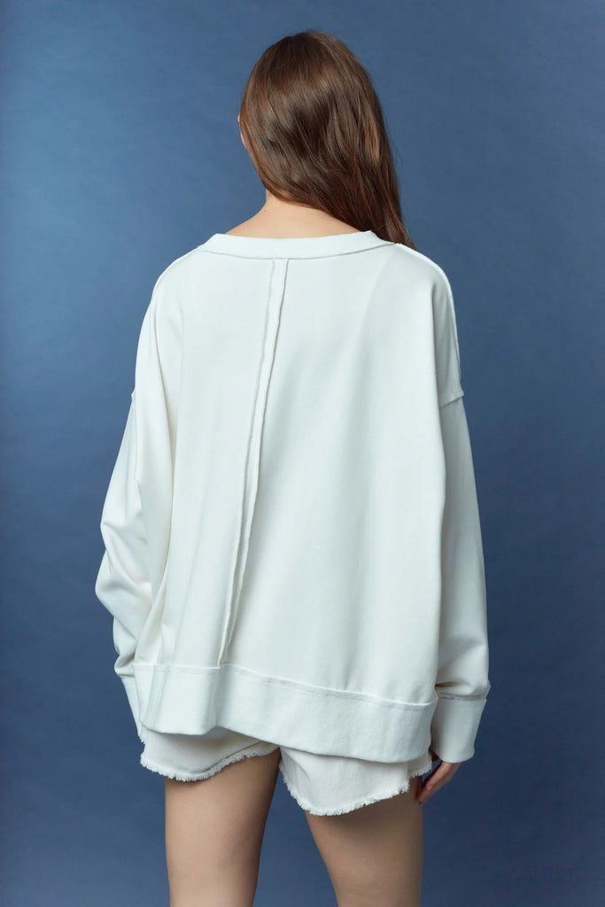 Woman wearing a white oversized sweatshirt with frayed appliqué letters spelling “TOUCHDOWN,” with the O’s replaced by a football and helmet, styled with white frayed shorts against a blue background.