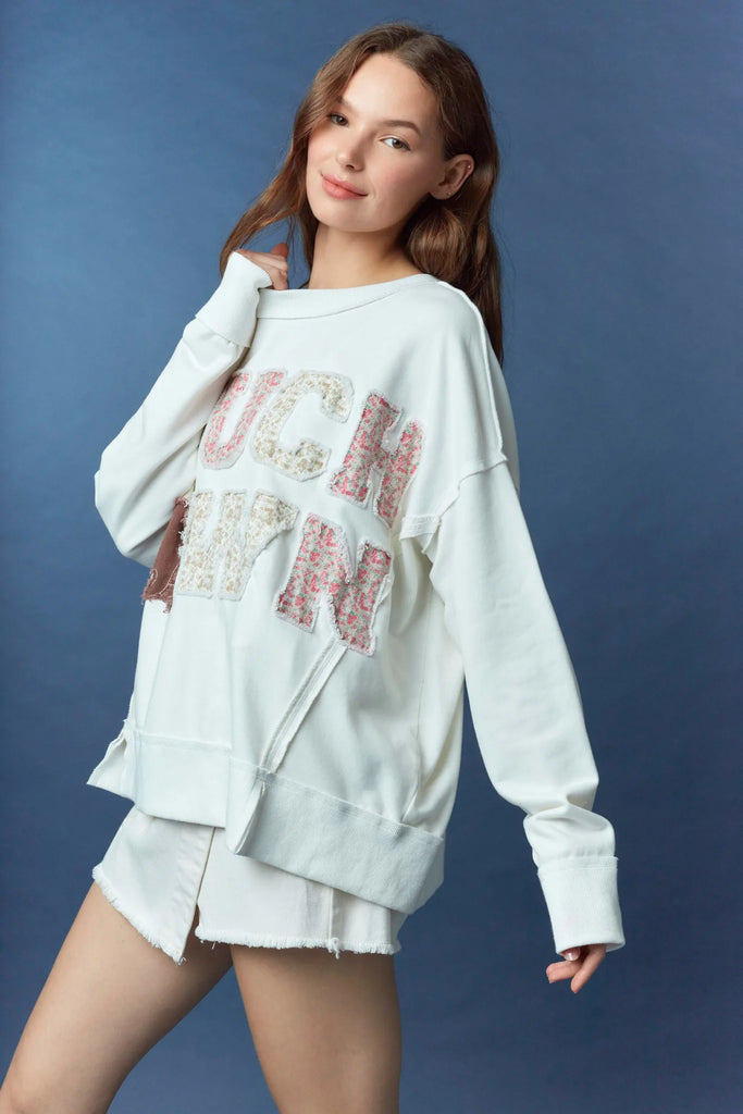 Woman wearing a white oversized sweatshirt with frayed appliqué letters spelling “TOUCHDOWN,” with the O’s replaced by a football and helmet, styled with white frayed shorts against a blue background.