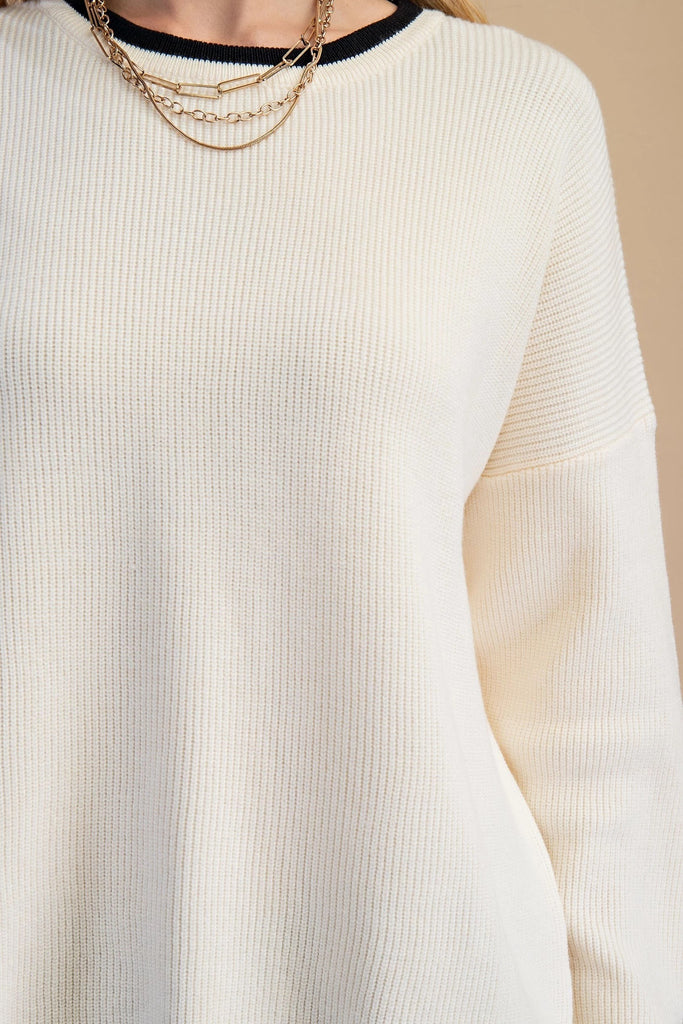An ivory knit sweater featuring a black contrasting trim on the crew neckline and a distinctive wavy black hem.