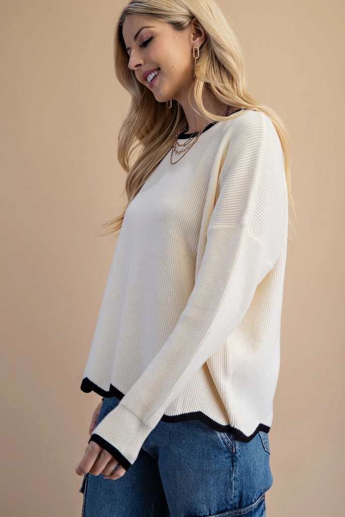 An ivory knit sweater featuring a black contrasting trim on the crew neckline and a distinctive wavy black hem.