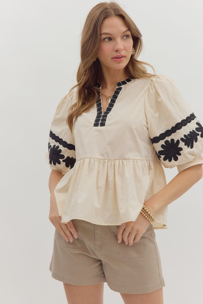 A woman wears a light-colored woven top with three-quarter length bubble sleeves and a peplum ruffle hem, featuring black floral embroidery on the sleeves and neck. She is wearing it with light brown shorts.