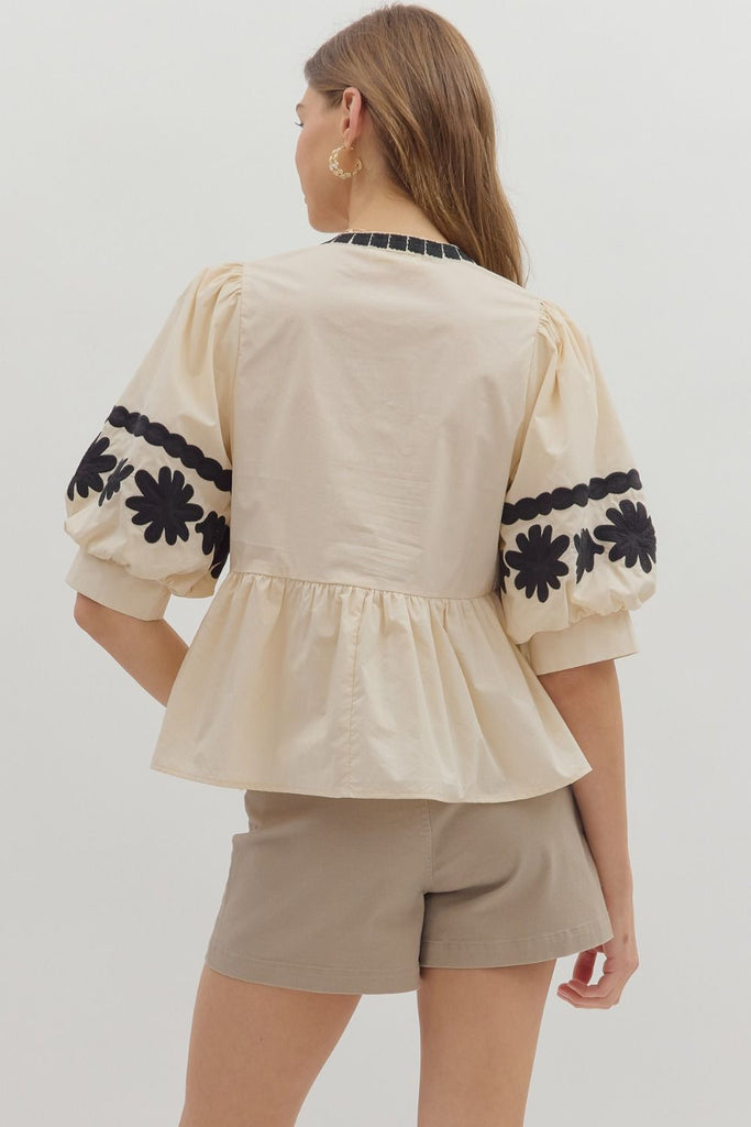 A woman wears a light-colored woven top with three-quarter length bubble sleeves and a peplum ruffle hem, featuring black floral embroidery on the sleeves and neck. She is wearing it with light brown shorts.