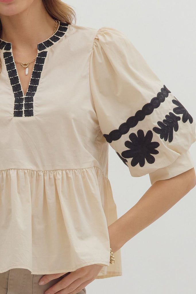 A woman wears a light-colored woven top with three-quarter length bubble sleeves and a peplum ruffle hem, featuring black floral embroidery on the sleeves and neck. She is wearing it with light brown shorts.