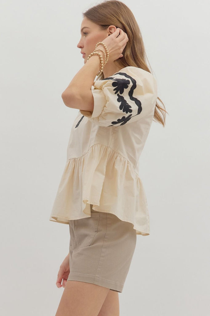 A woman wears a light-colored woven top with three-quarter length bubble sleeves and a peplum ruffle hem, featuring black floral embroidery on the sleeves and neck. She is wearing it with light brown shorts.