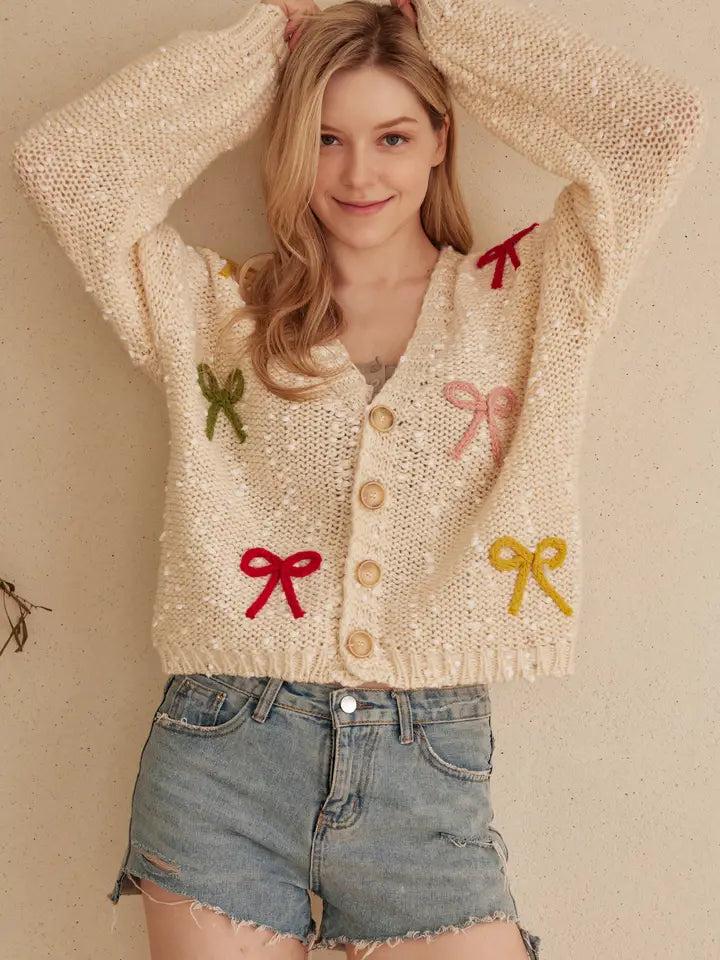 A woman wears a cream-colored, chunky knit cardigan with colorful ribbon bows in various colors attached to the front. The cardigan has a V-neckline and button front closure.