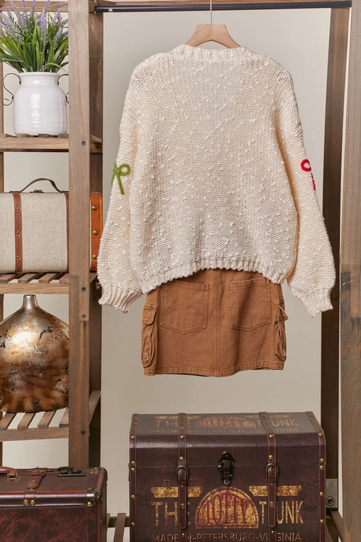 A woman wears a cream-colored, chunky knit cardigan with colorful ribbon bows in various colors attached to the front. The cardigan has a V-neckline and button front closure.