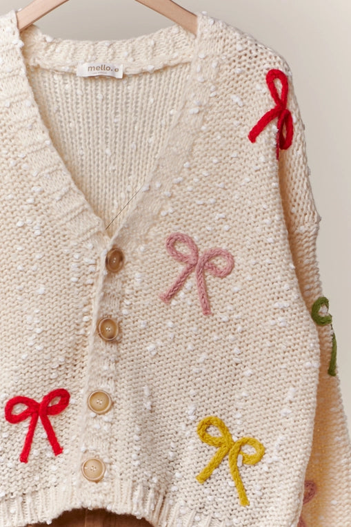 A woman wears a cream-colored, chunky knit cardigan with colorful ribbon bows in various colors attached to the front. The cardigan has a V-neckline and button front closure.