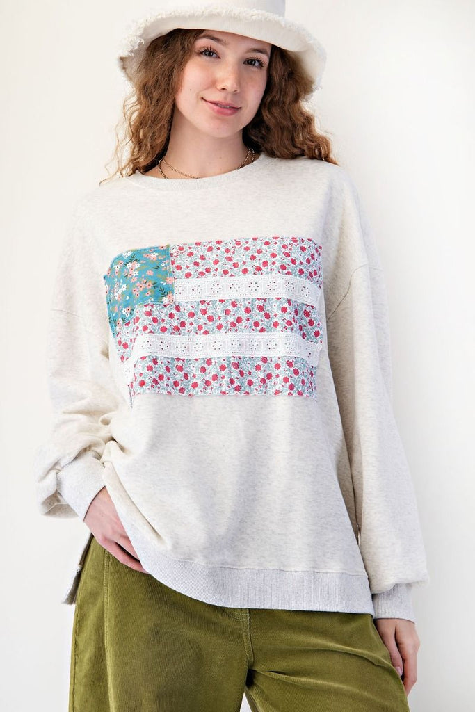 Woman wearing a light heather gray oversized terry pullover featuring a patchwork American flag design on the front, with floral print panels and eyelet lace stripes. Styled with olive green corduroy pants and a soft white bucket hat for a cozy, casual look