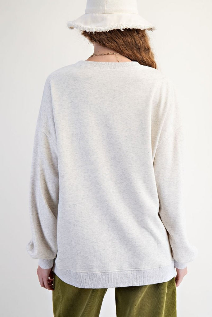 Woman wearing a light heather gray oversized terry pullover featuring a patchwork American flag design on the front, with floral print panels and eyelet lace stripes. Styled with olive green corduroy pants and a soft white bucket hat for a cozy, casual look