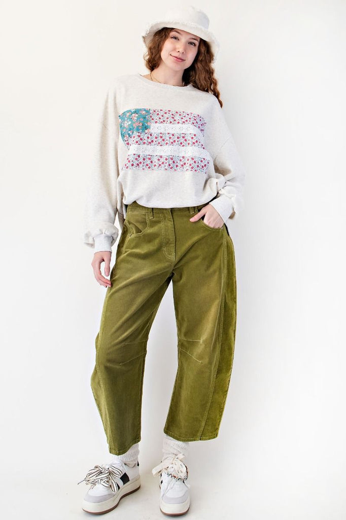 Woman wearing a light heather gray oversized terry pullover featuring a patchwork American flag design on the front, with floral print panels and eyelet lace stripes. Styled with olive green corduroy pants and a soft white bucket hat for a cozy, casual look