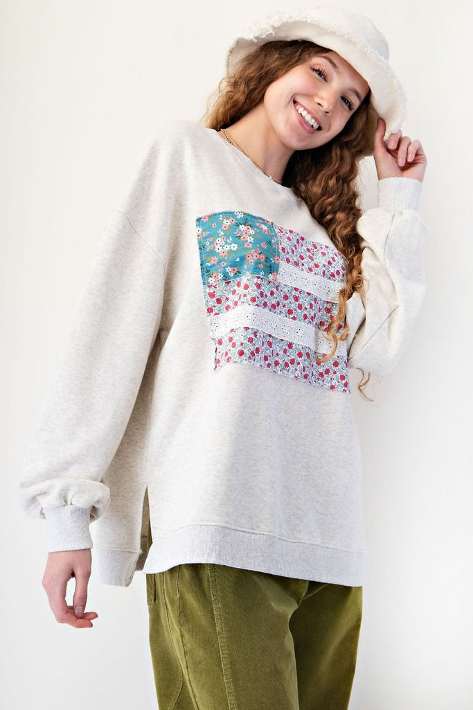 Woman wearing a light heather gray oversized terry pullover featuring a patchwork American flag design on the front, with floral print panels and eyelet lace stripes. Styled with olive green corduroy pants and a soft white bucket hat for a cozy, casual look
