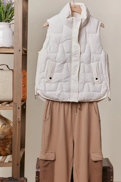 An ivory-colored, quilted puffer vest with a high mock neck is shown hanging, layered over a pair of brown cargo-style pants. The vest has a front snap closure and side pockets.