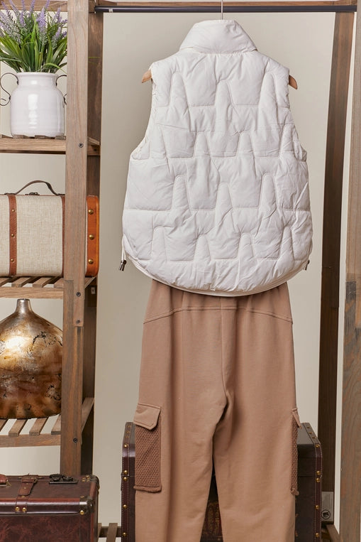An ivory-colored, quilted puffer vest with a high mock neck is shown hanging, layered over a pair of brown cargo-style pants. The vest has a front snap closure and side pockets.