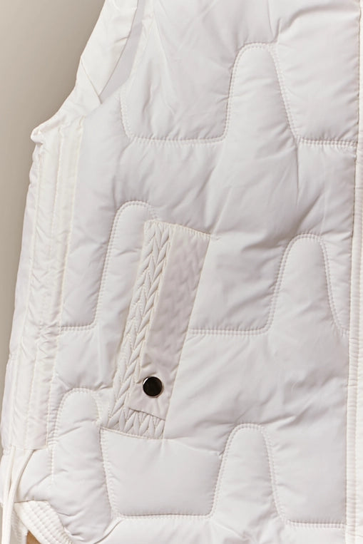 An ivory-colored, quilted puffer vest with a high mock neck is shown hanging, layered over a pair of brown cargo-style pants. The vest has a front snap closure and side pockets.
