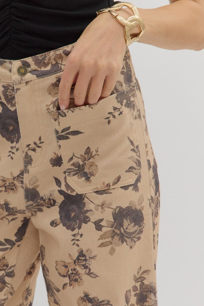 A person wears a pair of mid-rise, ankle-length, wide-leg pants with a dark floral print on a light tan background, styled with tan cowboy boots. The pants have front and back patch pockets.