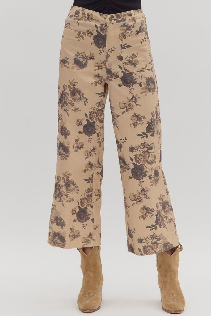 A person wears a pair of mid-rise, ankle-length, wide-leg pants with a dark floral print on a light tan background, styled with tan cowboy boots. The pants have front and back patch pockets.