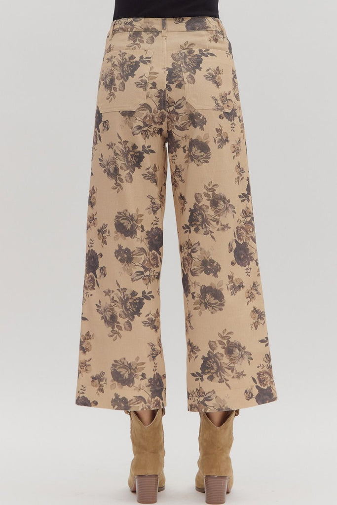 A person wears a pair of mid-rise, ankle-length, wide-leg pants with a dark floral print on a light tan background, styled with tan cowboy boots. The pants have front and back patch pockets.