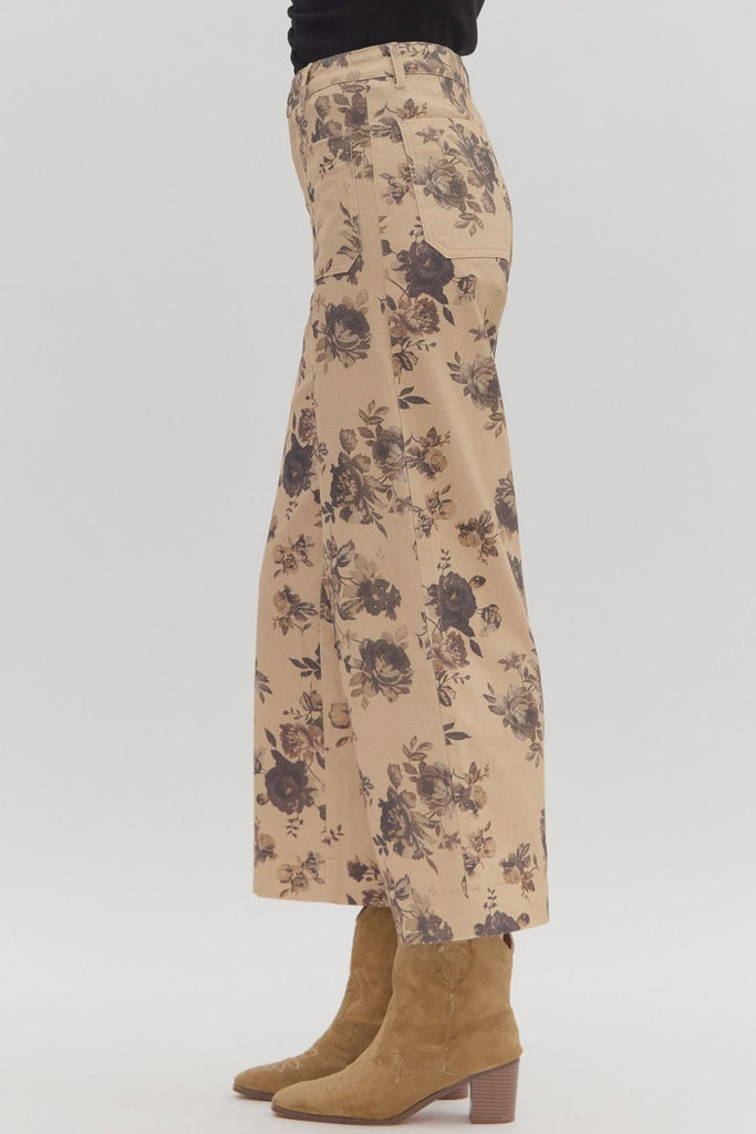 A person wears a pair of mid-rise, ankle-length, wide-leg pants with a dark floral print on a light tan background, styled with tan cowboy boots. The pants have front and back patch pockets.