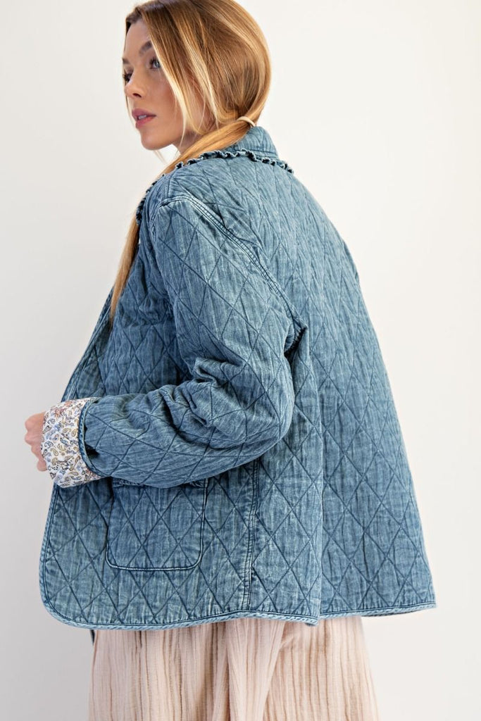 A washed denim jacket with a quilted diamond pattern. It has a bow tie-front closure, a ruffled collar and cuffs, and large front pockets with ruffled trim.