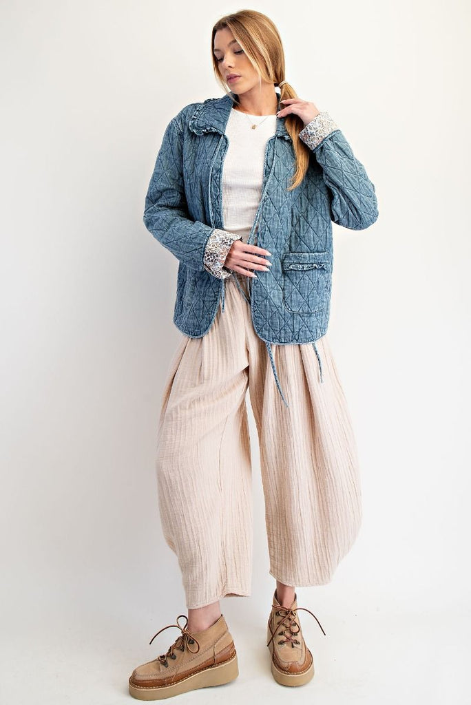 A washed denim jacket with a quilted diamond pattern. It has a bow tie-front closure, a ruffled collar and cuffs, and large front pockets with ruffled trim.