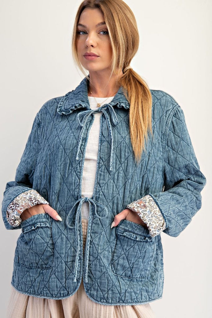 A washed denim jacket with a quilted diamond pattern. It has a bow tie-front closure, a ruffled collar and cuffs, and large front pockets with ruffled trim.