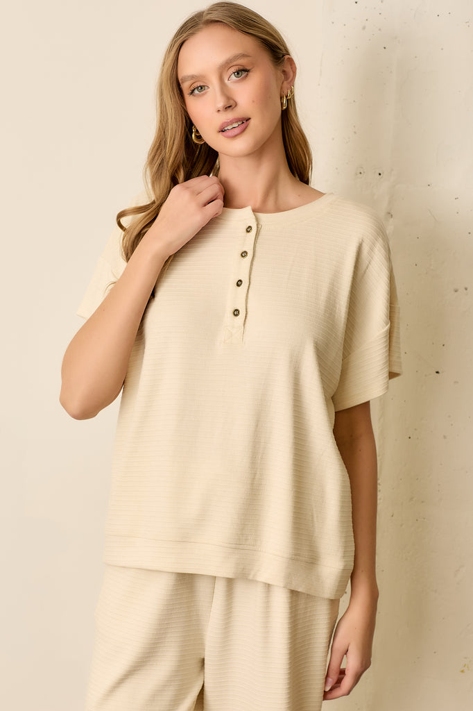Beige ribbed short sleeve top with vertical stripe texture, round neckline, and functional front buttons. Soft knit fabric with a relaxed fit.