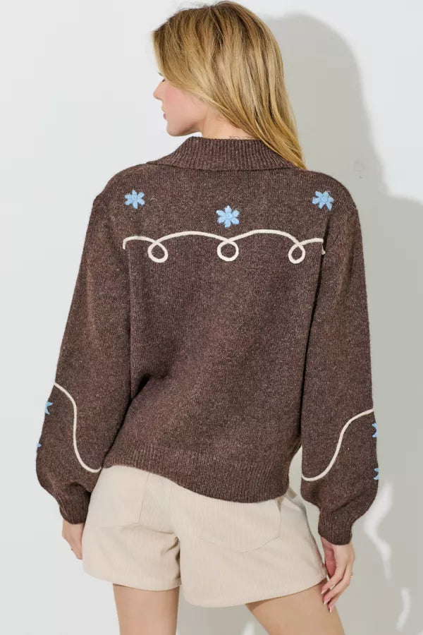 A brown sweater knit cardigan with a white embroidered swirl and line pattern and light blue embroidered floral accents. It has a collar and a button-up front with large pearlescent buttons.