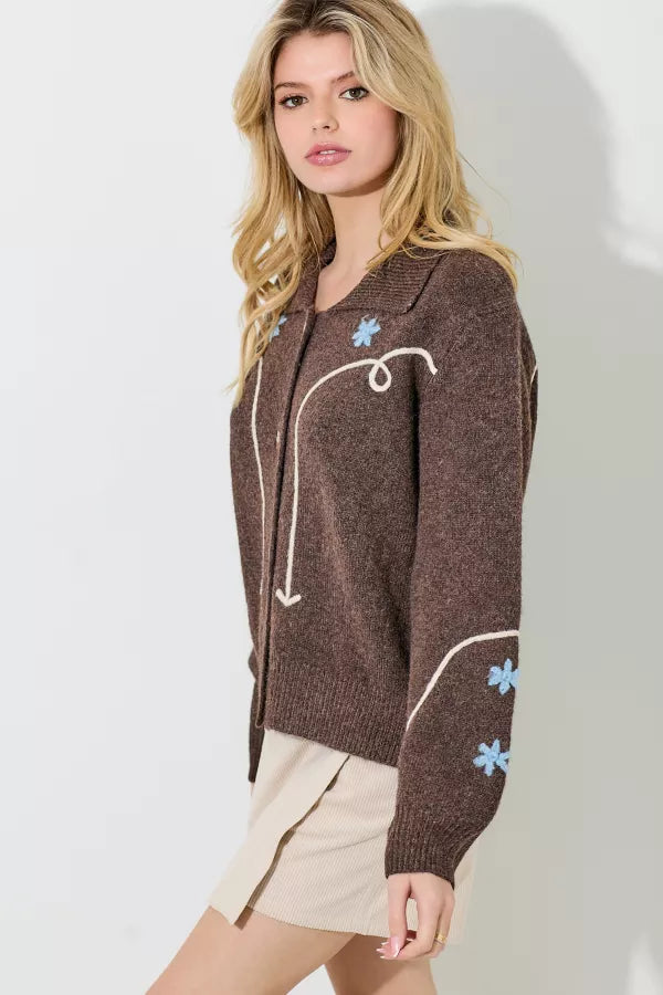 A brown sweater knit cardigan with a white embroidered swirl and line pattern and light blue embroidered floral accents. It has a collar and a button-up front with large pearlescent buttons.
