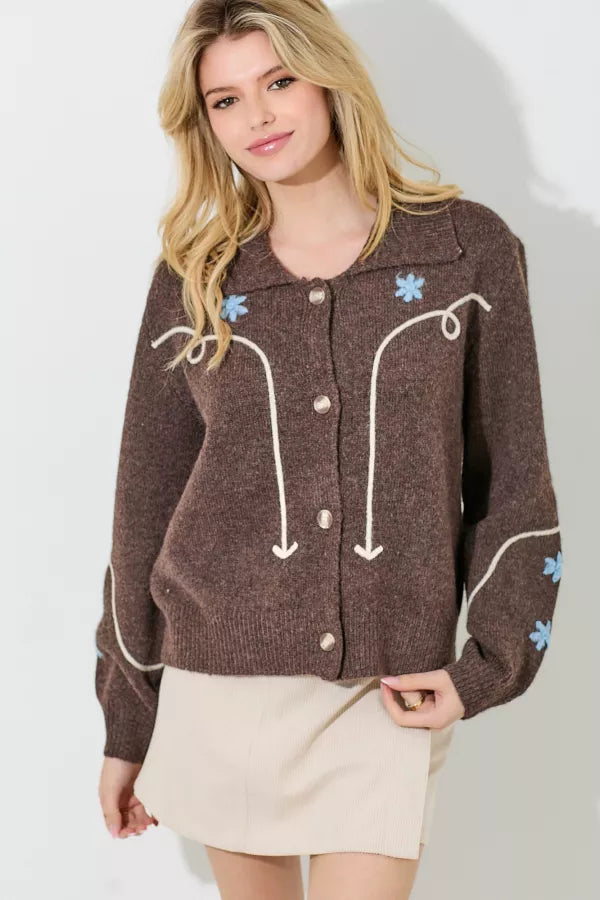 A brown sweater knit cardigan with a white embroidered swirl and line pattern and light blue embroidered floral accents. It has a collar and a button-up front with large pearlescent buttons.
