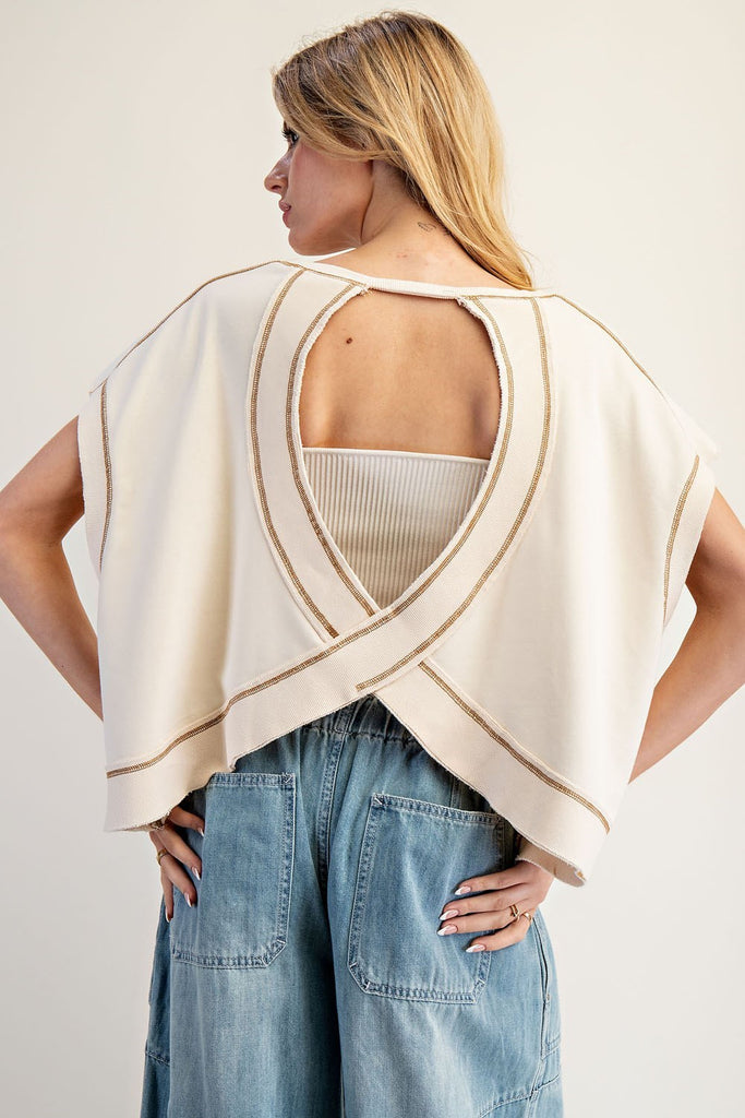 Model wearing a cream-colored sleeveless top with a wide, draped open-back design featuring a crisscross hem and contrast stitching. The top is layered over a ribbed white bandeau and paired with high-waisted denim pants with large back pockets.