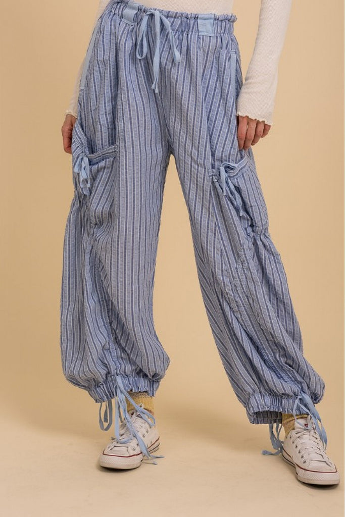 A model wears light blue and white striped parachute pants with an oversized, low-slung fit. The pants feature ruched drawstring detailing at the cuffs and pockets, with large side cargo pockets tied with matching fabric. Styled with a long-sleeve white top, white socks, and classic white Converse sneakers.
