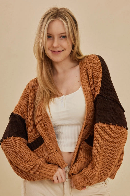 A woman wears a large, oversized cardigan with a color-block design in warm rust and dark brown. She is wearing it over a white crop top.