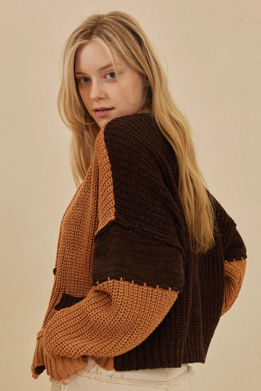 A woman wears a large, oversized cardigan with a color-block design in warm rust and dark brown. She is wearing it over a white crop top.