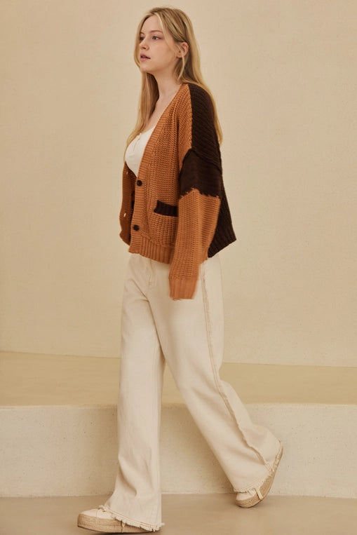 A woman wears a large, oversized cardigan with a color-block design in warm rust and dark brown. She is wearing it over a white crop top.