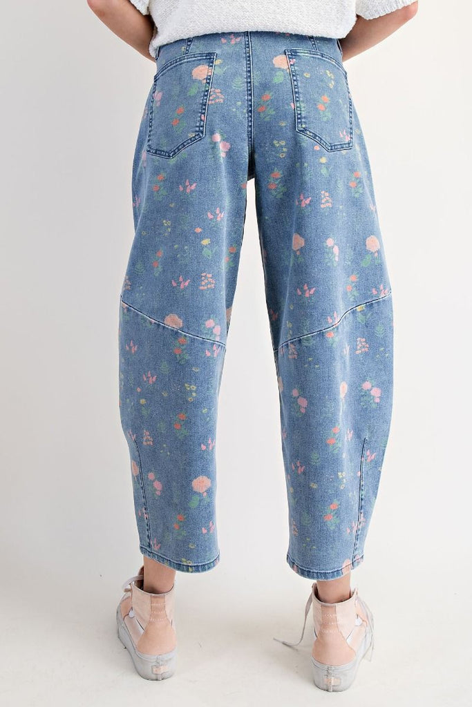 A pair of washed denim barrel-leg pants with a floral print in pink, peach, and green. The pants are high-waisted and cropped at the ankle.