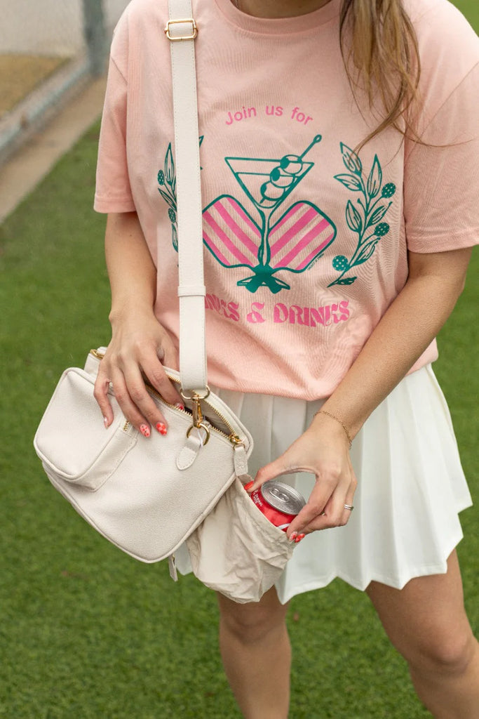 Person wearing a pink t-shirt with graphics and text, holding a white bag and a can on grass.