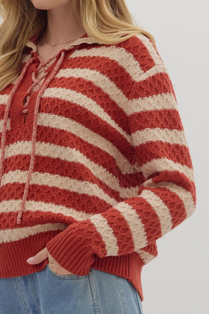 A woman models a striped, long-sleeve sweater in red and cream with a lace-up front and a collar. She is wearing it with light wash jeans.