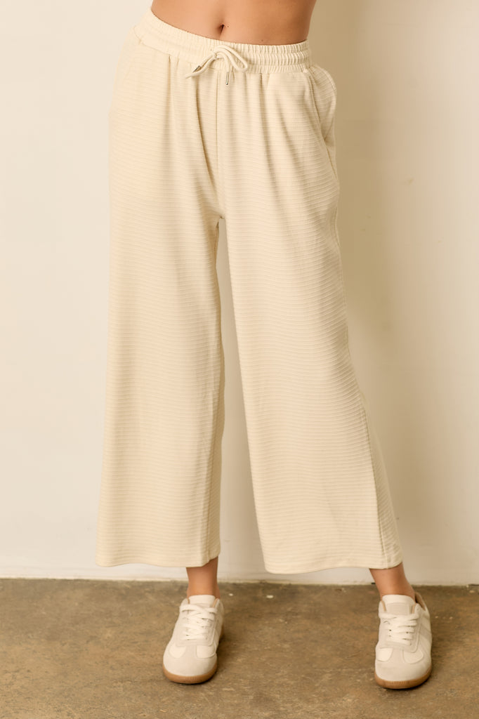 Beige ribbed knit pants with vertical stripes, elastic waistband, drawstring front, and side pockets. Relaxed fit with soft stretch fabric.