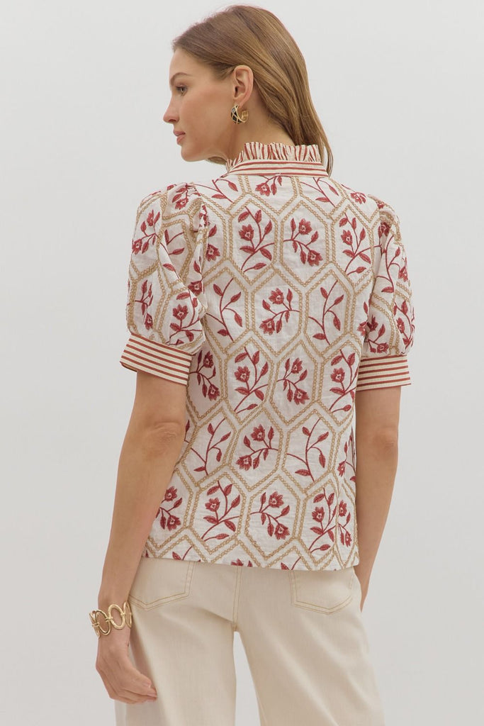A white top with a brick-red floral and gold chain-link embroidered pattern. It has short puff sleeves, a high ruffled neck, and a front button placket with a contrasting red and white striped pattern.