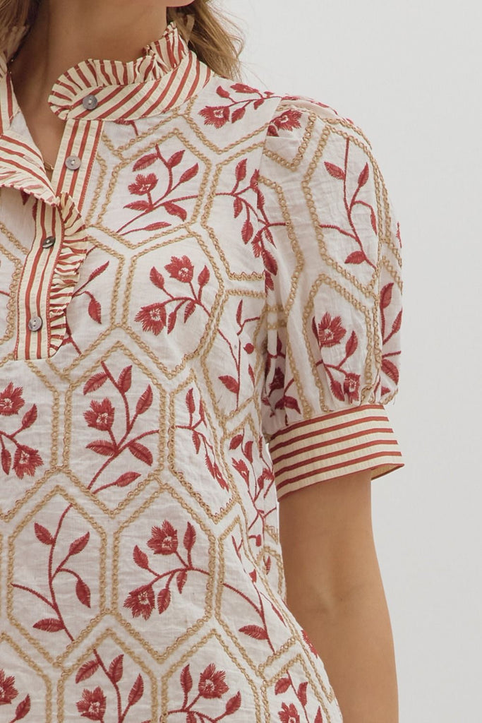 A white top with a brick-red floral and gold chain-link embroidered pattern. It has short puff sleeves, a high ruffled neck, and a front button placket with a contrasting red and white striped pattern.