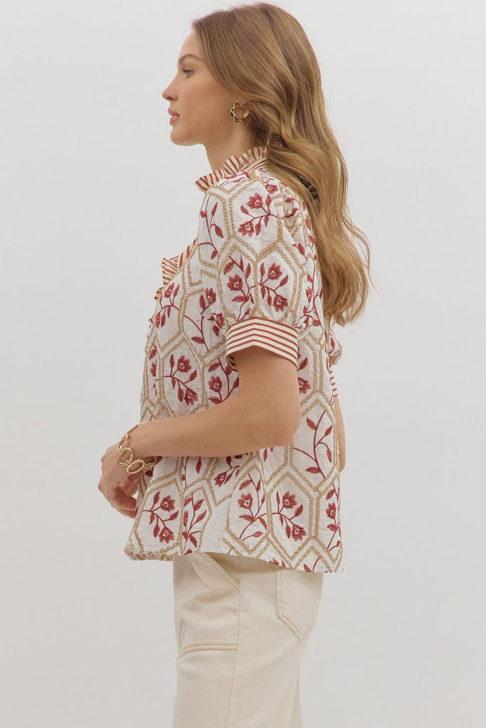 A white top with a brick-red floral and gold chain-link embroidered pattern. It has short puff sleeves, a high ruffled neck, and a front button placket with a contrasting red and white striped pattern.