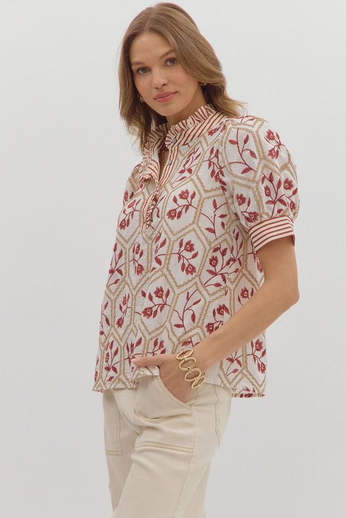 A white top with a brick-red floral and gold chain-link embroidered pattern. It has short puff sleeves, a high ruffled neck, and a front button placket with a contrasting red and white striped pattern.