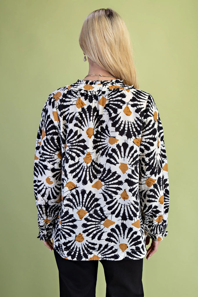 A woman wearing a vibrant, black, white, and gold tie-dye inspired top. The long-sleeved blouse has a v-neck with a tie detail ending in black tassels, and is paired with black pants. The background is a soft, solid green color.