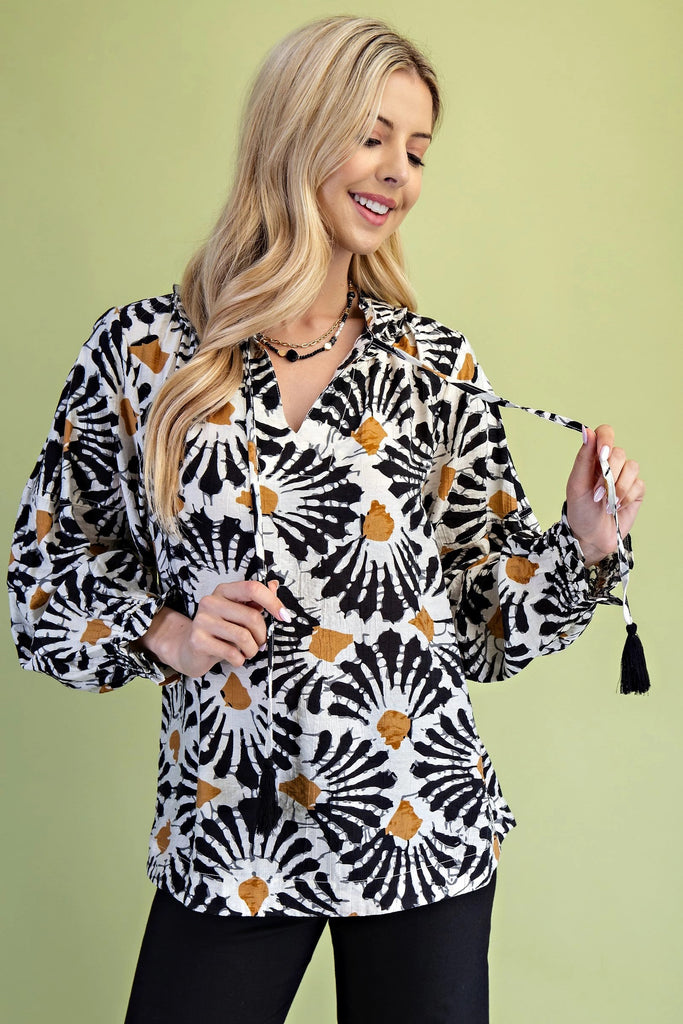 A product shot featuring a smiling blonde woman wearing a vibrant, black, white, and gold tie-dye inspired top. The long-sleeved blouse has a v-neck with a tie detail ending in black tassels, and is paired with black pants. The background is a soft, solid green color.