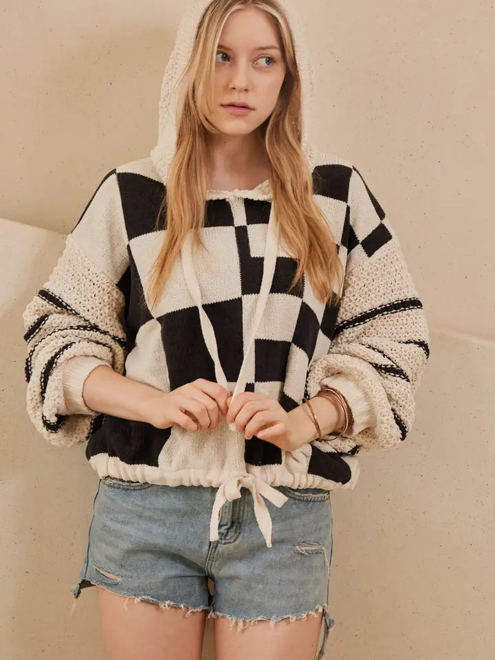 A woman models a hooded, black and off-white checkerboard sweater with textured, contrasting knit sleeves and a drawstring hem, styled with denim shorts.