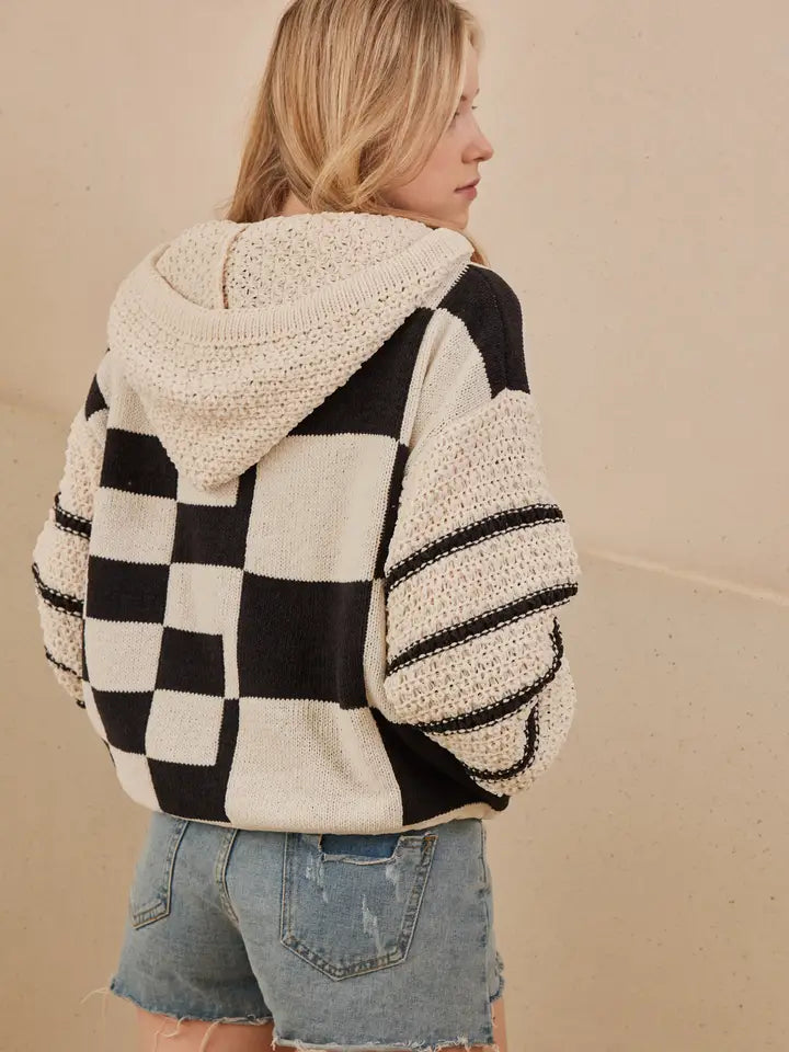 A woman models a hooded, black and off-white checkerboard sweater with textured, contrasting knit sleeves and a drawstring hem, styled with denim shorts.