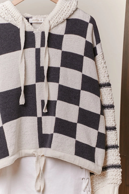 A woman models a hooded, black and off-white checkerboard sweater with textured, contrasting knit sleeves and a drawstring hem, styled with denim shorts.