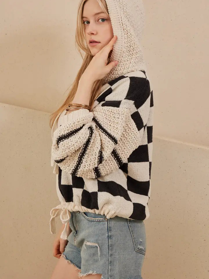 A woman models a hooded, black and off-white checkerboard sweater with textured, contrasting knit sleeves and a drawstring hem, styled with denim shorts.